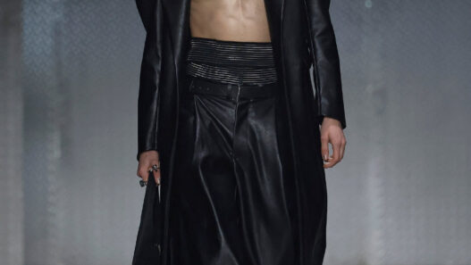 Male model walking runway in a long black leather coat open to reveal bare chest and black wide-leg pants, glossy slicked-back hair.