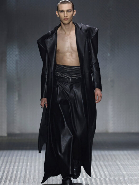 Male model walking runway in a long black leather coat open to reveal bare chest and black wide-leg pants, glossy slicked-back hair.