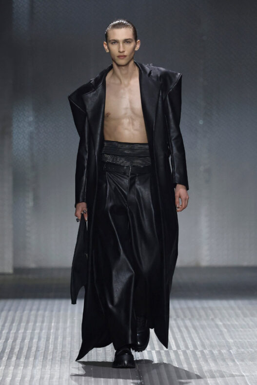 Male model walking runway in a long black leather coat open to reveal bare chest and black wide-leg pants, glossy slicked-back hair.