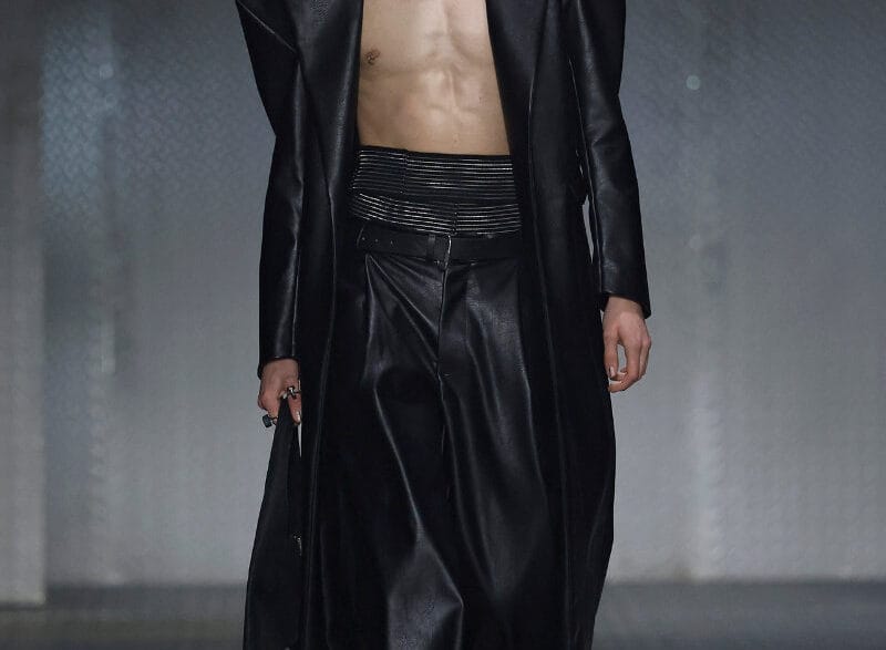 Male model walking runway in a long black leather coat open to reveal bare chest and black wide-leg pants, glossy slicked-back hair.