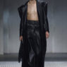 Male model walking runway in a long black leather coat open to reveal bare chest and black wide-leg pants, glossy slicked-back hair.