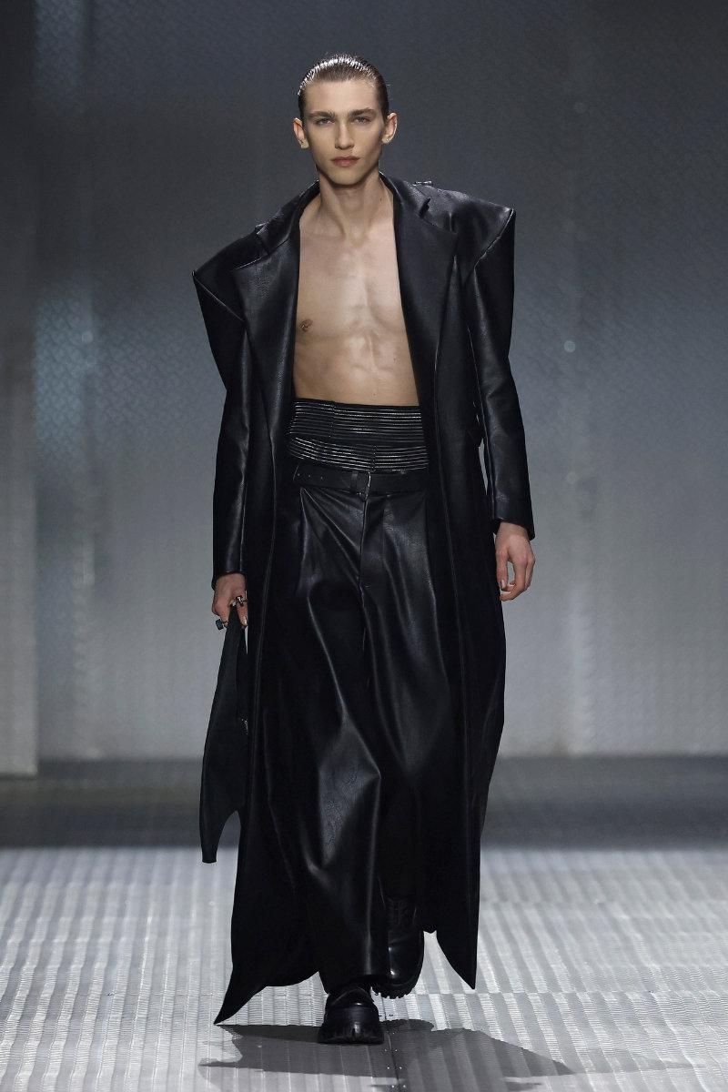 Male model walking runway in a long black leather coat open to reveal bare chest and black wide-leg pants, glossy slicked-back hair.