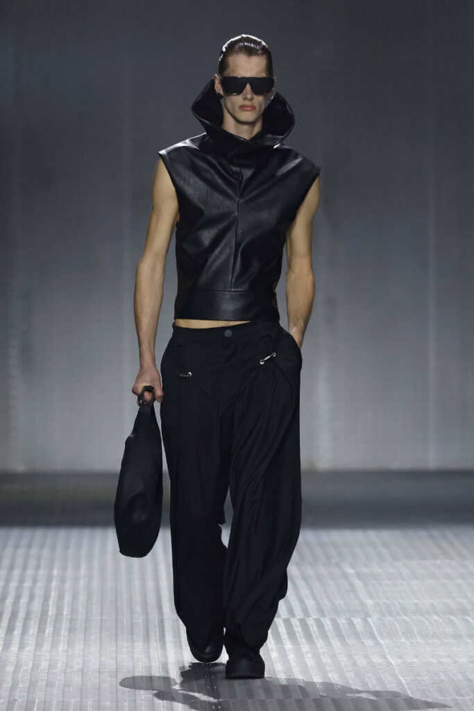 Model on a runway wearing an all-black outfit: sleeveless leather high-collar top, loose black pants, oversized sunglasses, and a black cap held in hand.