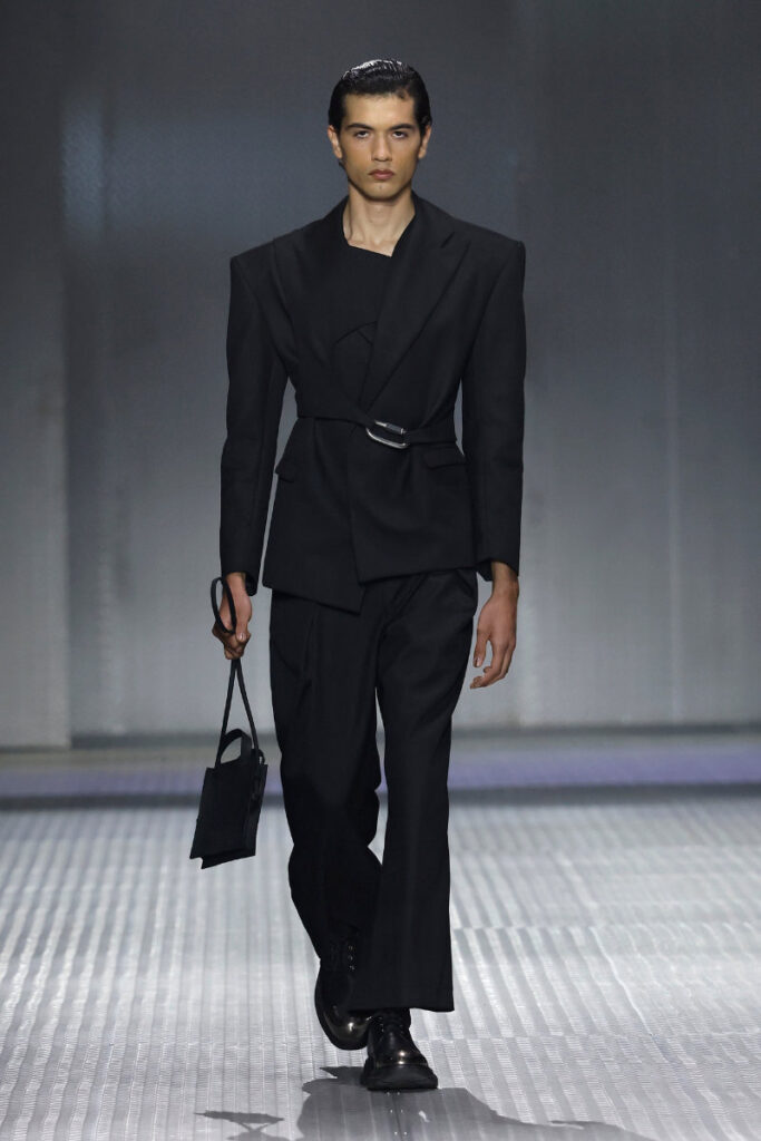 Male model on a runway wearing a black tailored blazer with belt and wide-leg pants, holding a small black handbag.