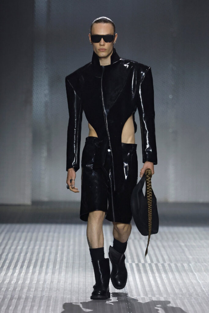 Model on a runway in a glossy black, cut‑out coat with matching shorts and oversized sunglasses.