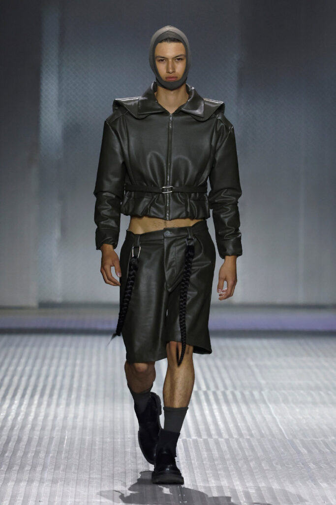 Model on a runway wearing a black leather cropped jacket with a waist belt, matching knee-length shorts, a grey hood, and long braided straps hanging from the pockets.