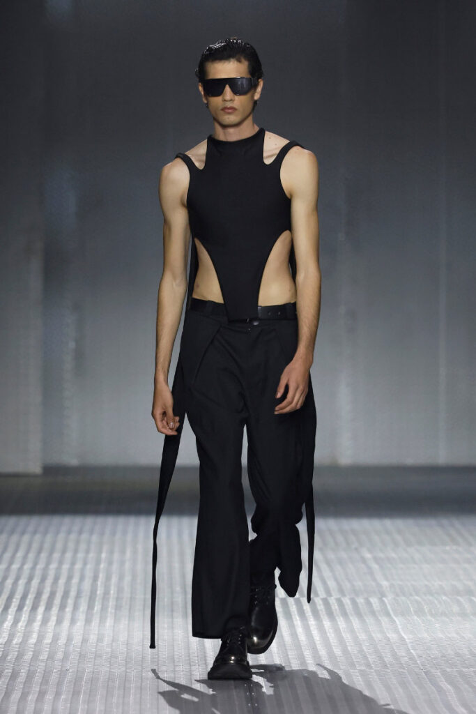 Male model on a runway wearing a black sleeveless top with side cutouts, belt, wide-leg pants, and dark sunglasses.