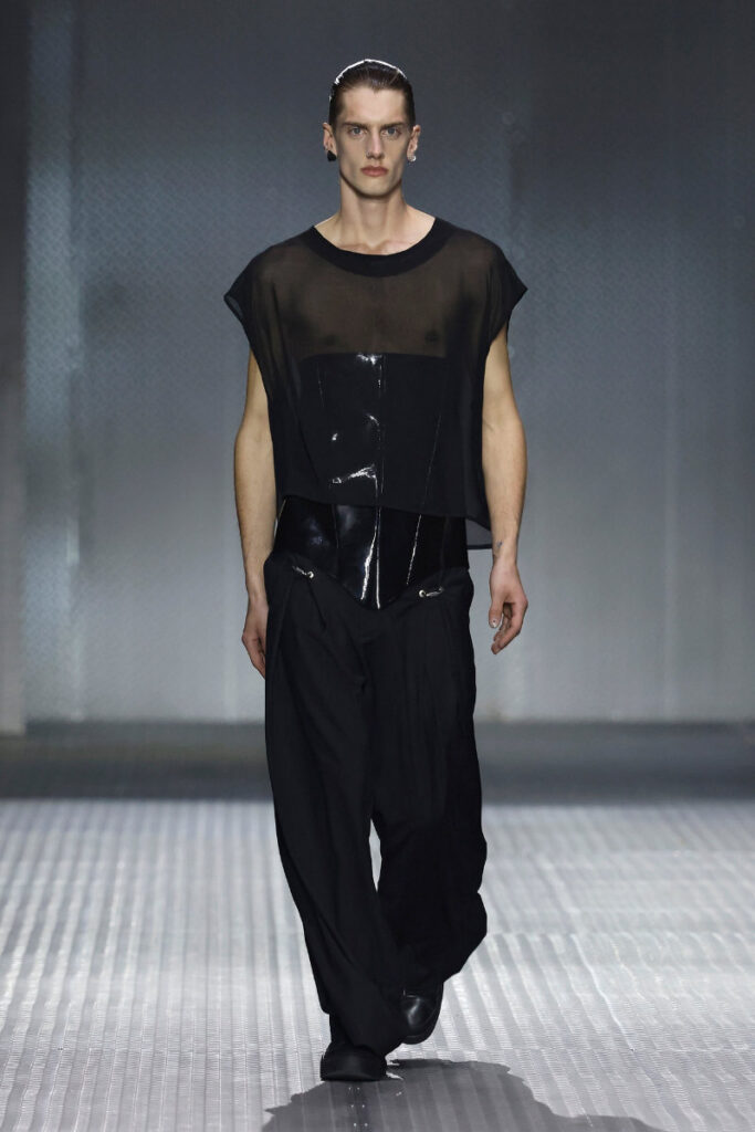 Male fashion model walking the runway in a sheer black top layered over a glossy black corset and wide black trousers.