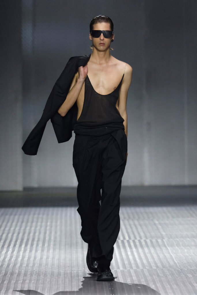 Model walking a runway in an all-black outfit: sheer sleeveless top, loose pants, jacket draped over the shoulder, and dark sunglasses.