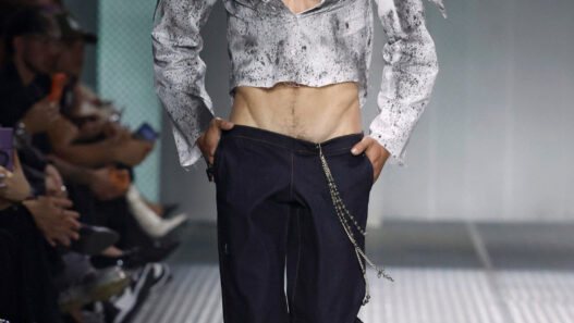 Male model on a runway wearing a metallic cropped gray jacket, open chest, dark trousers with a silver chain accessory.