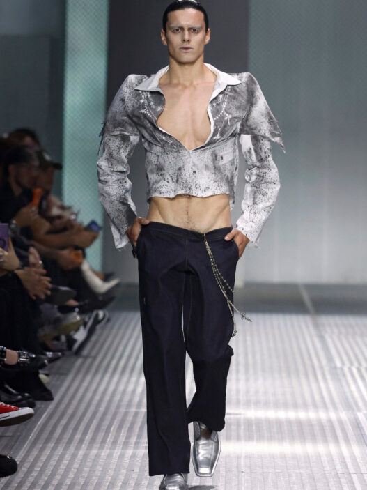Male model on a runway wearing a metallic cropped gray jacket, open chest, dark trousers with a silver chain accessory.