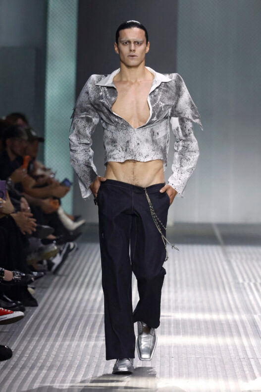 Male model on a runway wearing a metallic cropped gray jacket, open chest, dark trousers with a silver chain accessory.
