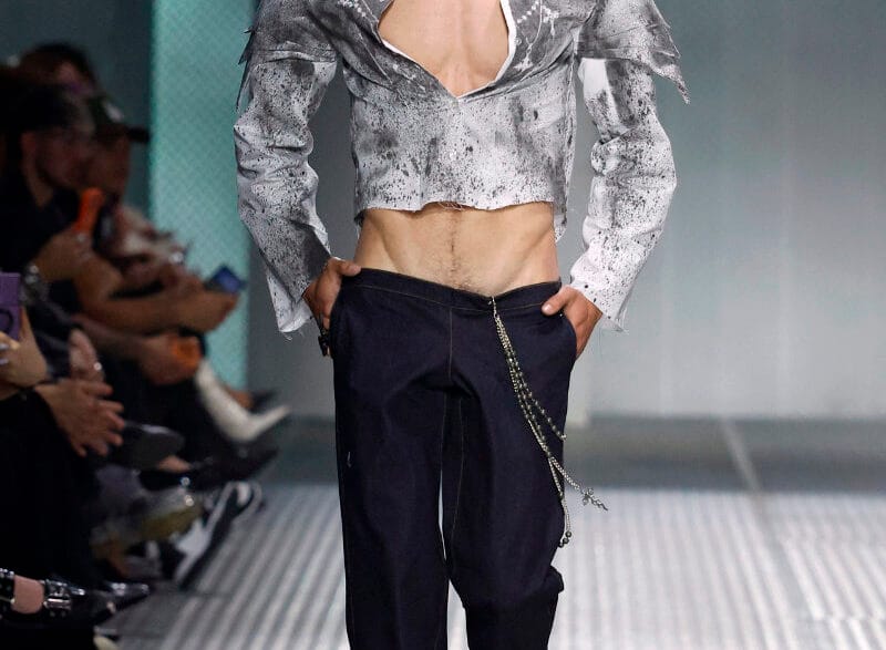 Male model on a runway wearing a metallic cropped gray jacket, open chest, dark trousers with a silver chain accessory.