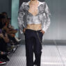 Male model on a runway wearing a metallic cropped gray jacket, open chest, dark trousers with a silver chain accessory.