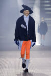 Male model walks the runway in an oversized navy coat over a light blue shirt, bright orange pants, gray socks, black loafers, blue gloves, and a wide black hat.