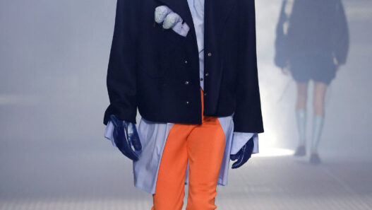 Male model walks the runway in an oversized navy coat over a light blue shirt, bright orange pants, gray socks, black loafers, blue gloves, and a wide black hat.