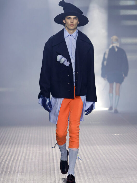 Male model walks the runway in an oversized navy coat over a light blue shirt, bright orange pants, gray socks, black loafers, blue gloves, and a wide black hat.