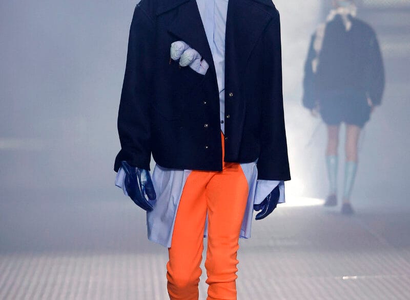 Male model walks the runway in an oversized navy coat over a light blue shirt, bright orange pants, gray socks, black loafers, blue gloves, and a wide black hat.