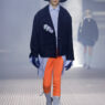Male model walks the runway in an oversized navy coat over a light blue shirt, bright orange pants, gray socks, black loafers, blue gloves, and a wide black hat.
