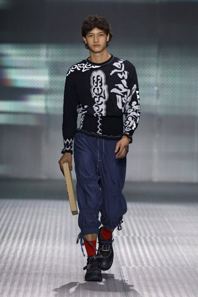 Male model on a fashion show runway, wearing a black white graphic sweater, navy cargo pants, red socks, buckled shoes, and holding a book.