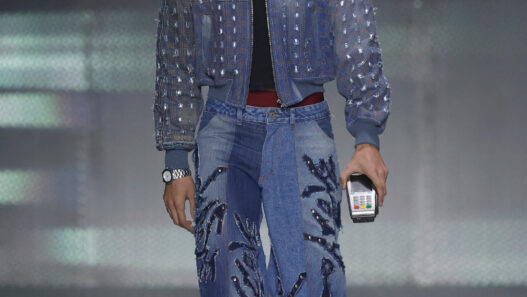 Model walking the runway in a blue denim outfit: studded jacket and embroidered jeans, holding a small handheld device in one hand.