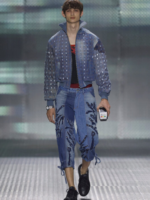 Model walking the runway in a blue denim outfit: studded jacket and embroidered jeans, holding a small handheld device in one hand.