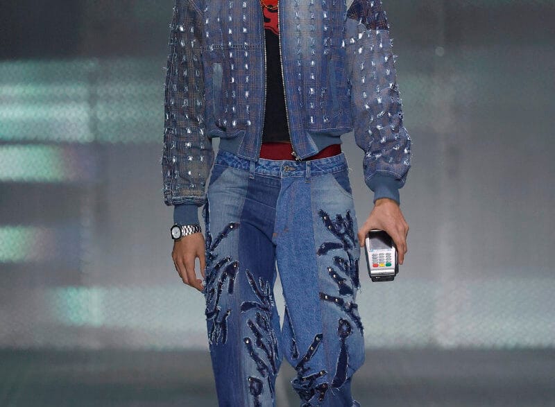 Model walking the runway in a blue denim outfit: studded jacket and embroidered jeans, holding a small handheld device in one hand.
