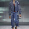 Model walking the runway in a blue denim outfit: studded jacket and embroidered jeans, holding a small handheld device in one hand.