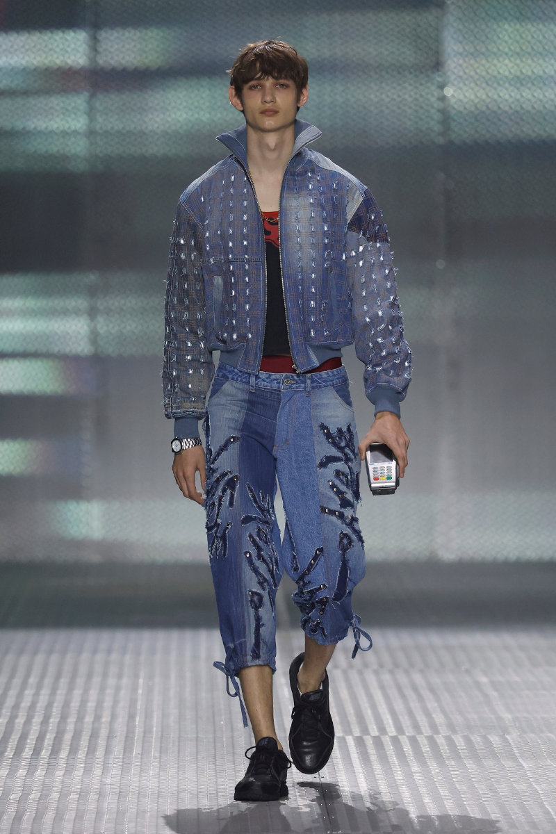 Model walking the runway in a blue denim outfit: studded jacket and embroidered jeans, holding a small handheld device in one hand.