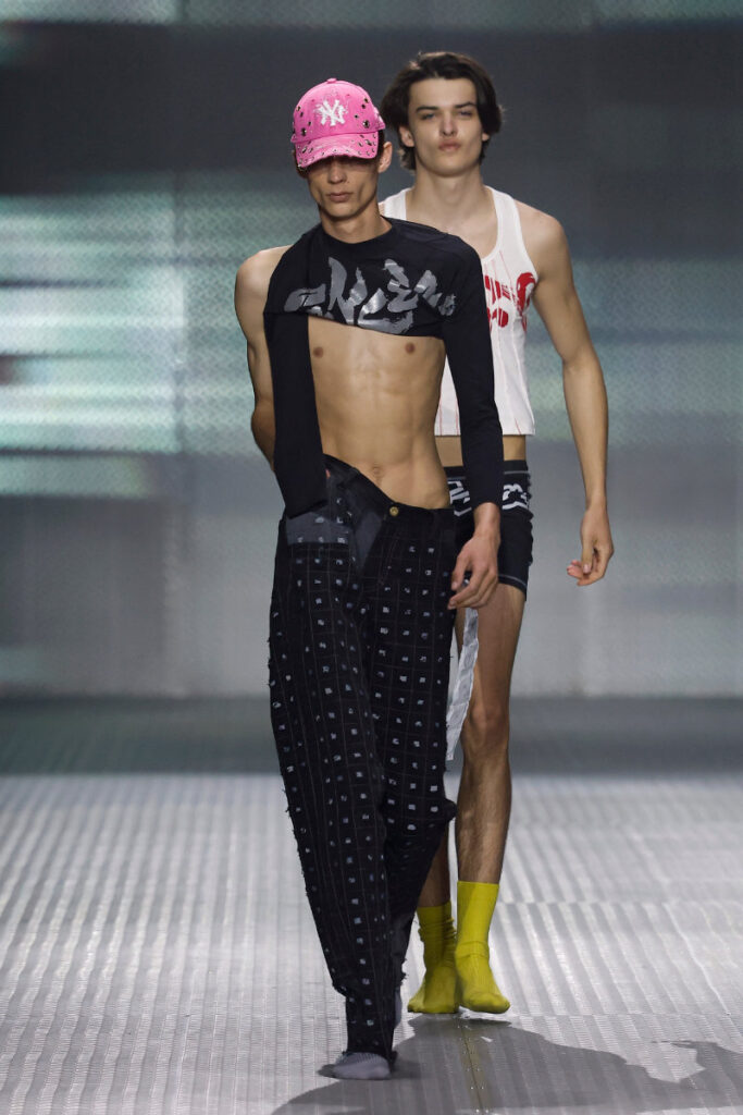 Two male models walk a runway; the front model wears a pink cap, black cropped top, and patterned pants showing his abs as the second model follows in a white sleeveless shirt and yellow socks.