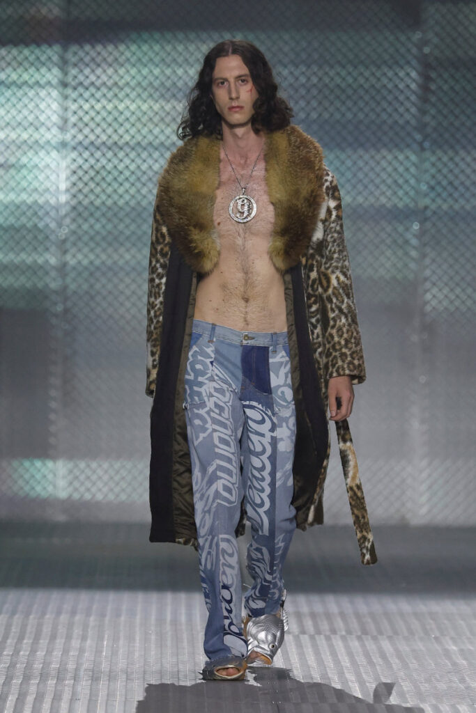 Male fashion model on a runway wearing an open leopard-print fur coat with a brown fur collar, bare chest, a large pendant necklace, and blue patterned jeans.