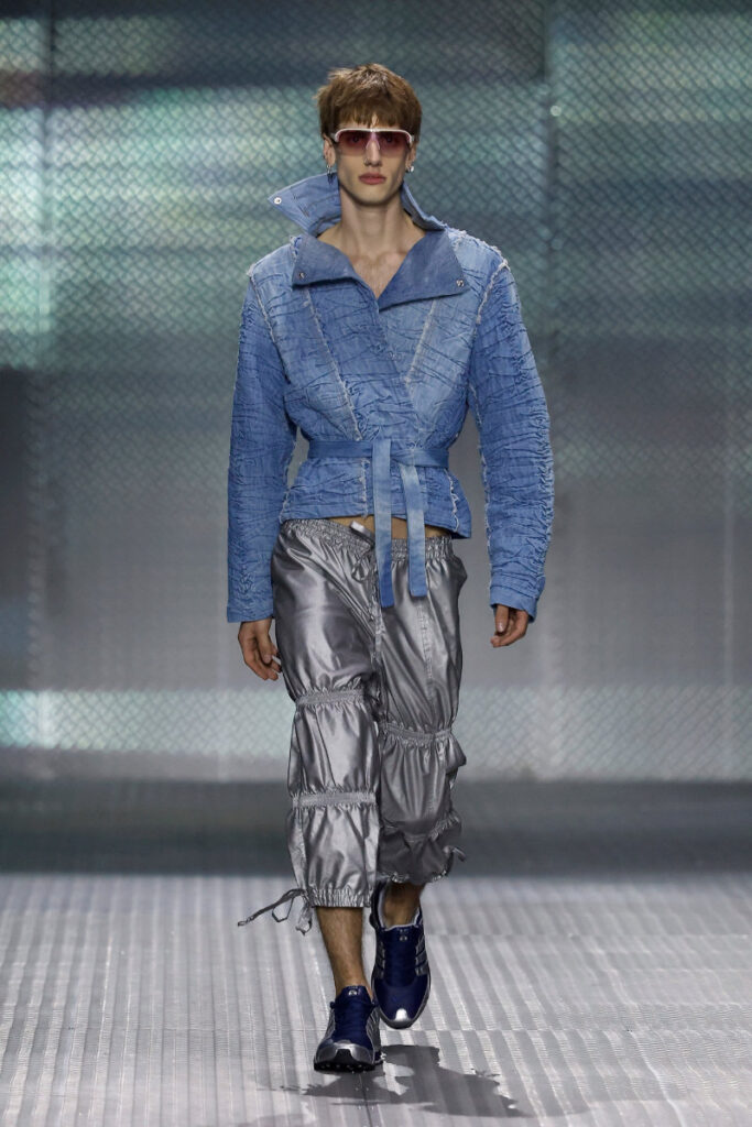 Male model walking a runway in a blue quilted jacket, belt, and metallic gray pants with blue sneakers and pink-tinted sunglasses