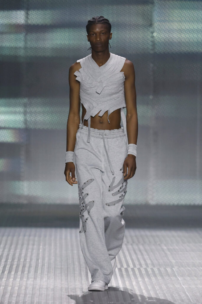 Male model on a runway wearing a distressed light gray sleeveless top and loose gray pants with metal rivet accents, walking a metallic catwalk.