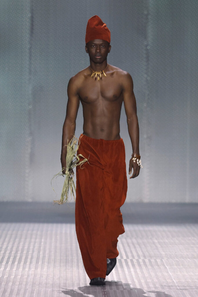Male fashion model on a runway wearing a red-orange wrap pants set with a matching head wrap, necklace, and holding dried grasses.