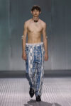 Male model walking a runway in blue, printed wide-leg pants, shirtless with a shell necklace and bracelets.