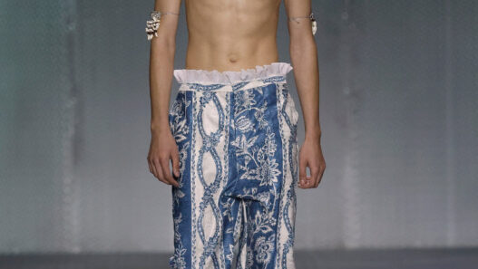 Male model walking a runway in blue, printed wide-leg pants, shirtless with a shell necklace and bracelets.