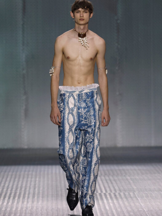 Male model walking a runway in blue, printed wide-leg pants, shirtless with a shell necklace and bracelets.