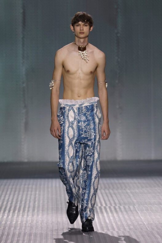 Male model walking a runway in blue, printed wide-leg pants, shirtless with a shell necklace and bracelets.