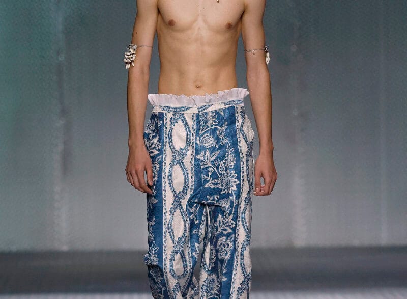 Male model walking a runway in blue, printed wide-leg pants, shirtless with a shell necklace and bracelets.