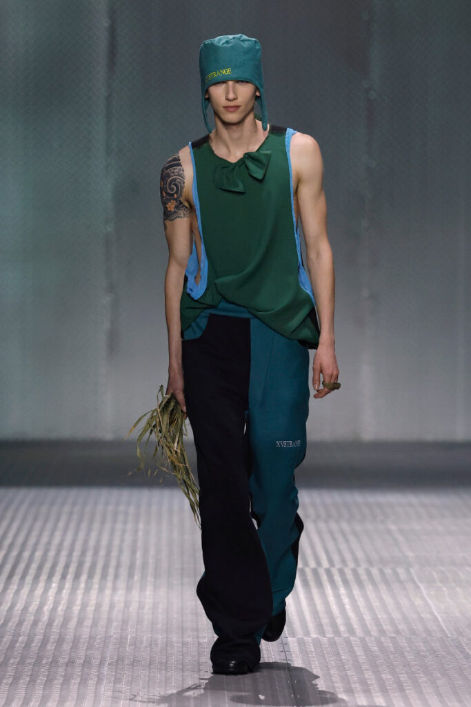 Male runway model walks the catwalk in a teal-green sleeveless top with blue side panels, matching teal hat, two-tone pants, and holding a tuft of grass.