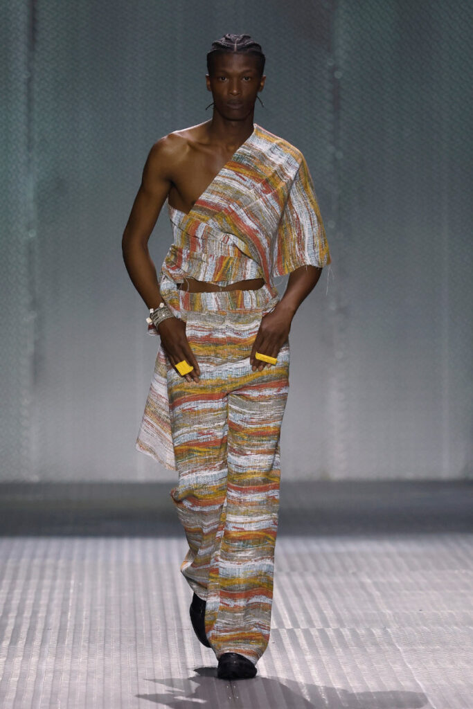 Male model walks the runway in a one-shoulder, striped two-piece outfit with wide-leg pants and yellow accessories.