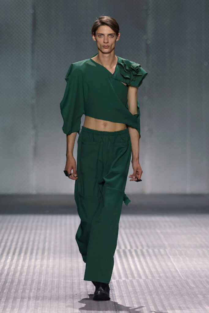 Model walking a fashion show runway in an emerald green cropped top with a one-shoulder puff sleeve and matching wide-leg pants