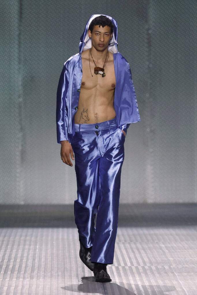 Male fashion model walks a runway wearing a shiny blue satin hooded jacket and matching pants, shirtless with a necklace and a wolf tattoo on his side.