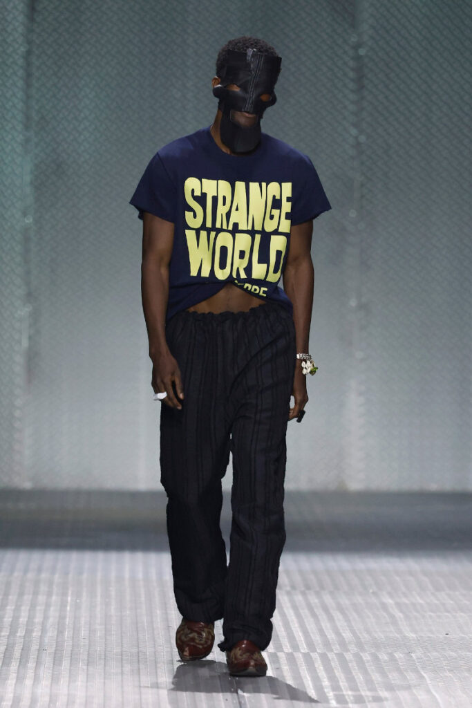 Male fashion model on a runway wearing a navy 'STRANGE WORLD' T‑shirt, black striped pants, and a black mask.