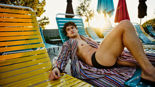 Man lounging shirtless in swim trunks on colorful poolside chairs at sunset, wearing a striped cover-up.