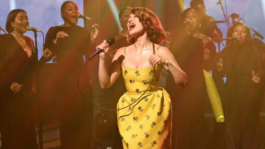 Female singer in a yellow floral dress performs center stage with a gospel choir behind her on a dimly lit stage.