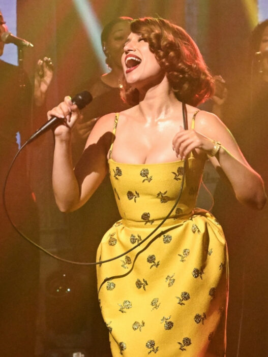 Female singer in a yellow floral dress performs center stage with a gospel choir behind her on a dimly lit stage.