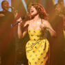 Female singer in a yellow floral dress performs center stage with a gospel choir behind her on a dimly lit stage.