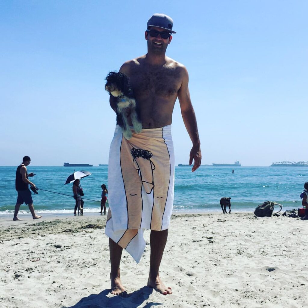 Man on a sunny beach wearing a cap and sunglasses, holding a small dog; a cartoon-pattern towel is wrapped around his waist.