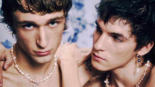 Two shirtless young men wearing pearl necklaces lean close, one with an arm around the other, against a blue-dyed backdrop.
